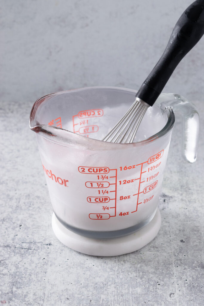 A chilled whisk just out of the freezer and a frosty 2-cup glass measuring cup with spout that has pink strawberry cream cold foam in it.