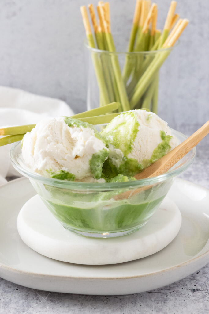 Scoops of vanilla ice cream floating in a bowl of matcha green tea with a spoon in the bowl and garnished with matcha green tea cream Pocky biscuit sticks.