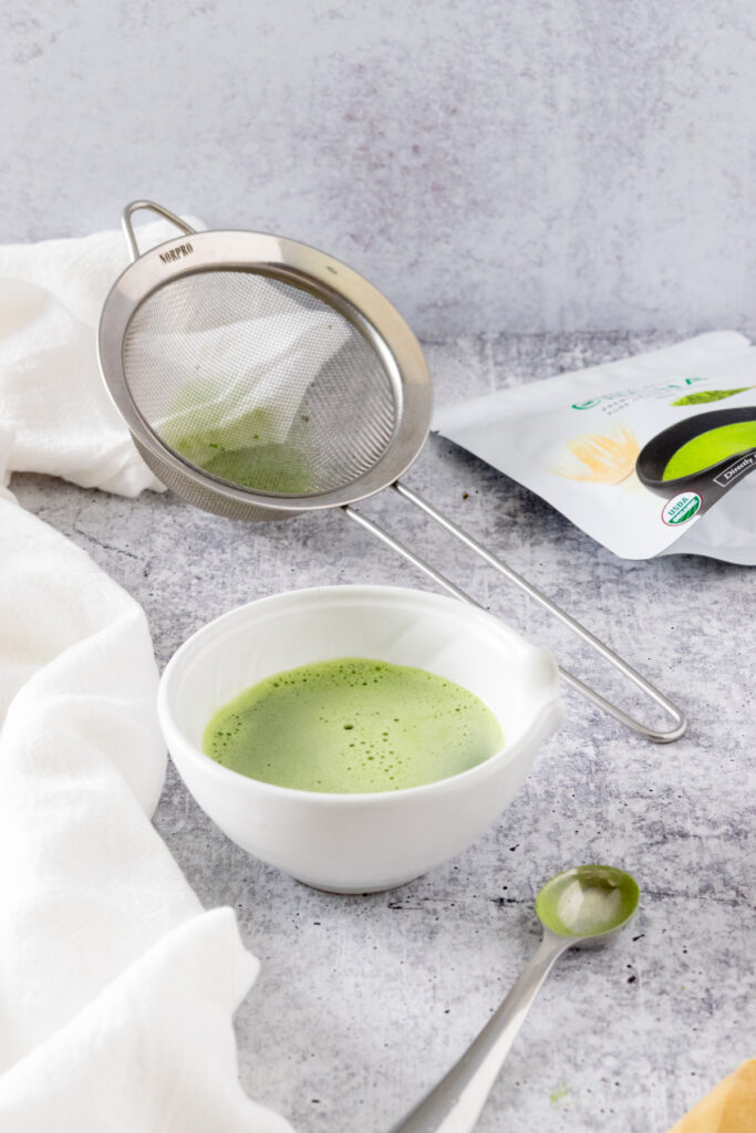 Small bowl of matcha that's been whisked in hot water sitting next to a bag of matcha powder and a strainer.