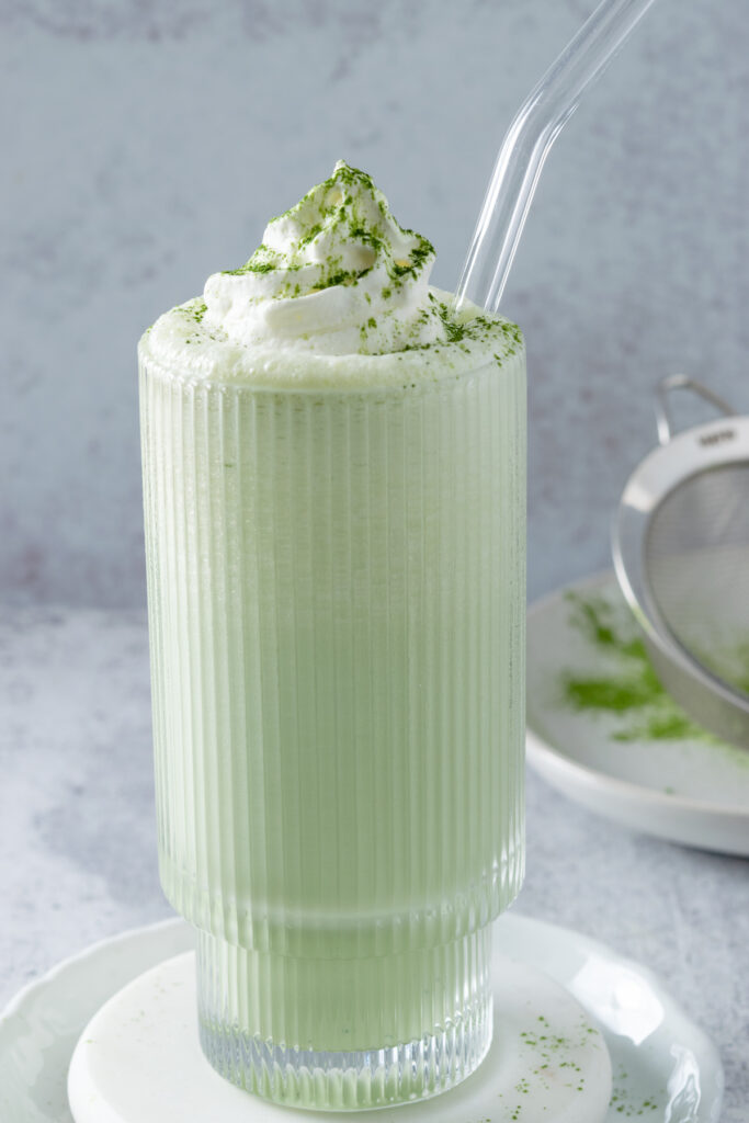 A matcha milkshake with whipped cream and matcha powder sprinkled on top. The milkshake is in a tall glass with glass straw.
