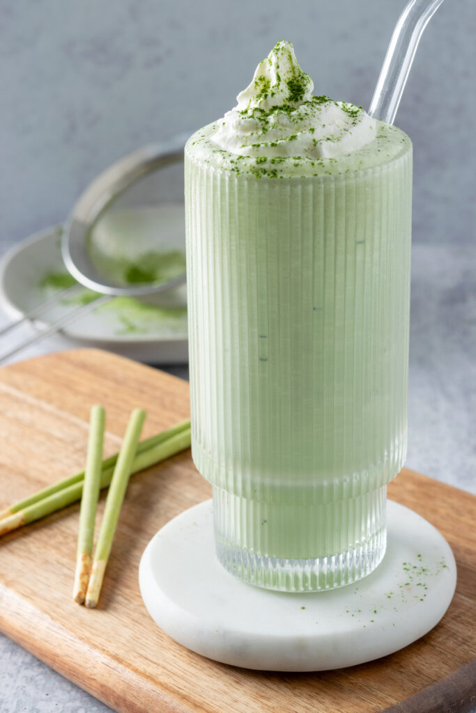 A homemade matcha milkshake in a glass on a white coaster. There are matcha flavored pocky biscuits next to the glass and a sifter with matcha powder on it in the background.