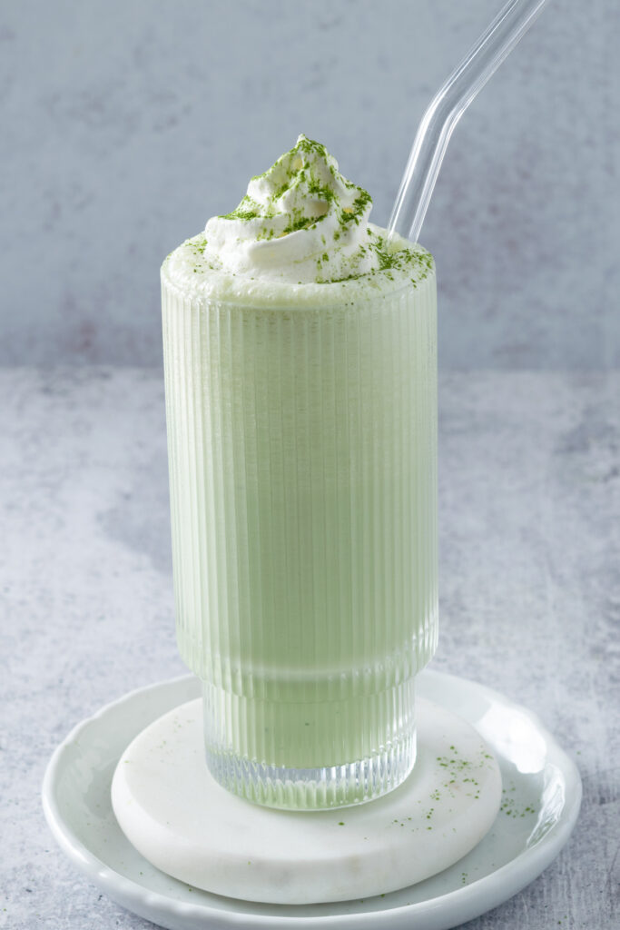 Matcha Milkshake in a tall glass with a glass straw in it and topped with whipped cream and a dusting of matcha green tea powder.