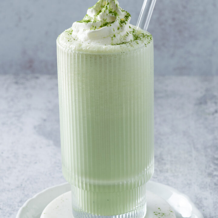 Matcha Milkshake in a tall glass with a glass straw in it and topped with whipped cream and a dusting of matcha green tea powder.