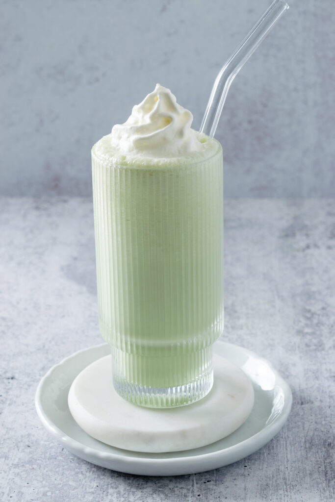 Homemade matcha milkshake in a tall, ribbed glass with glass straw on a white coaster. The milkshake is topped with whipped cream.