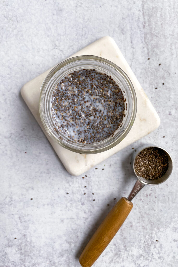Small bowl of chia seeds soaking in milk next to a 1 tablespoon measuring spoon that has some chia seeds in it.
