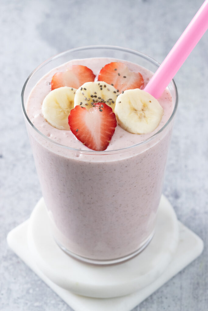 A top down view of a homemade strawberry banana smoothie with chia seeds. The smoothie is in a glass cup, has a pink straw in it, and is sitting on a white coaster. It's topped with a few slices of banana and strawberries and sprinkled with a few chia seeds.