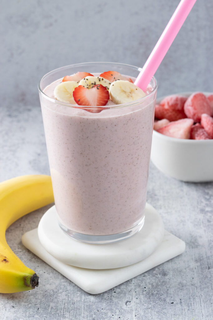 A strawberry banana smoothie in a glass cup which shows all the chia seeds in it. This pink smoothie is garnished with slices of banana and strawberries and has a straw in it. A whole banana and bowl of frozen strawberries are next to the smoothie.