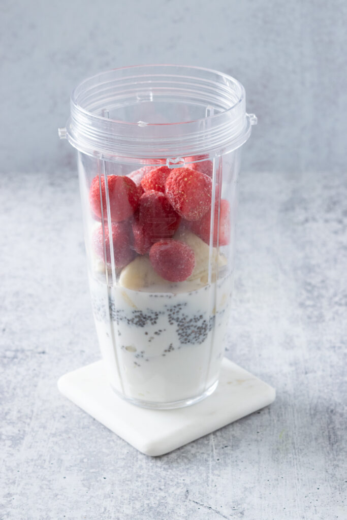 Frozen strawberries in a blender cup, along with ripe banana slices, milk, soaked chia seeds and yogurt.