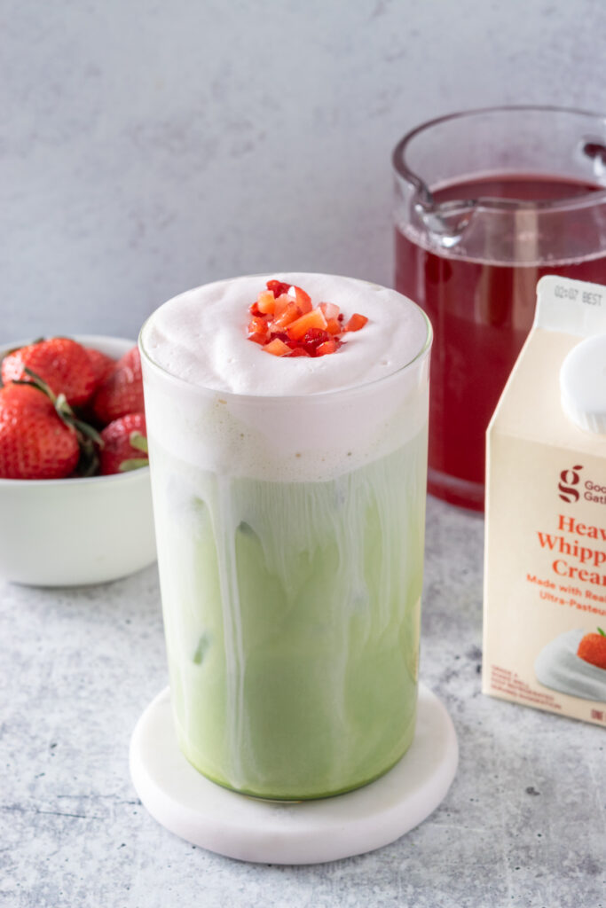 A creamy strawberry matcha latte garnished with small pile of chopped strawberries on top of the cold foam. The drink is in front of a carton of heavy whipping cream, bottle of strawberry syrup and bowl of strawberries.
