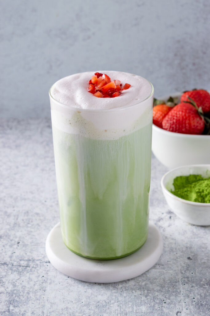 Pink and green layered iced matcha  latte topped with strawberry cream cold foam and garnished with chopped fresh strawberries. The drink is next to small bowls of strawberries and matcha green tea powder.