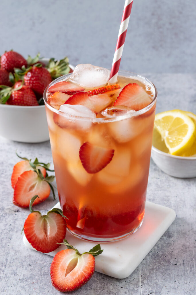 A refreshing glass of iced tea almost overflowing with ice and strawberry slices in it. There are two strawberries cut in half next to the iced tea glass, which is next to a bowl of strawberries and a smaller bowl of sliced lemons.