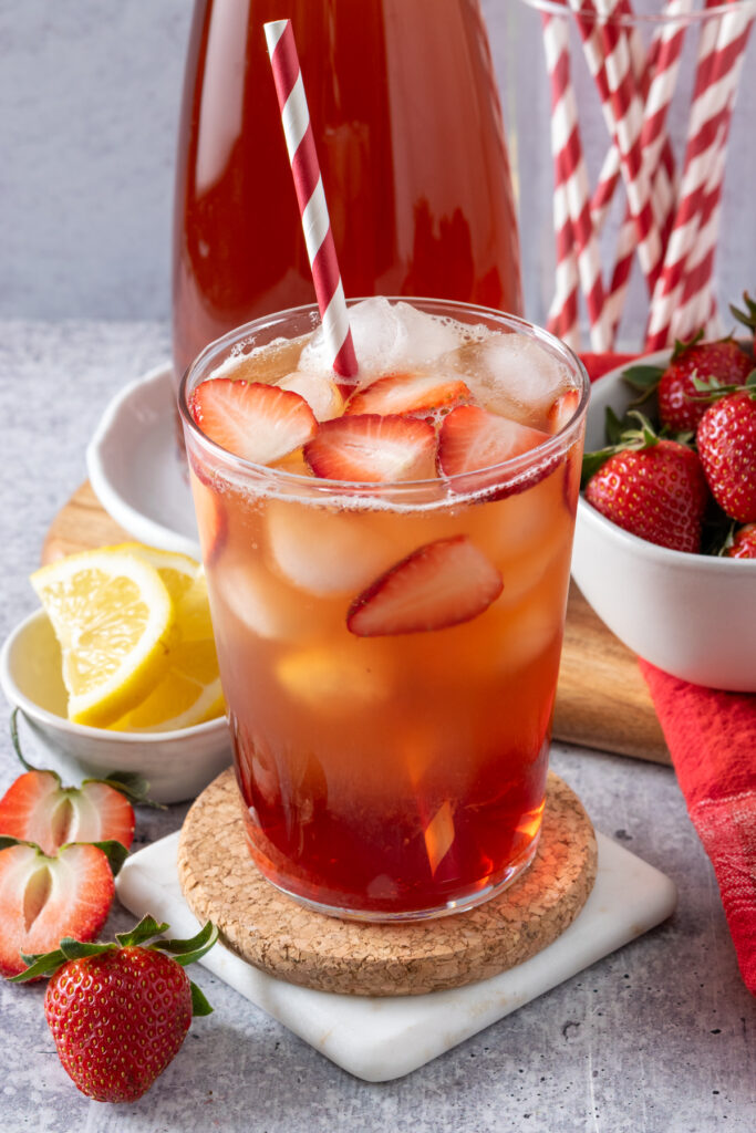 A frothy cup of strawberry iced tea with a red and white straw and ice and strawberry pieces in it. The cup is sitting in front of a pitcher of iced tea, bowl of sliced lemons, bowl of strawberries and container of more straws.