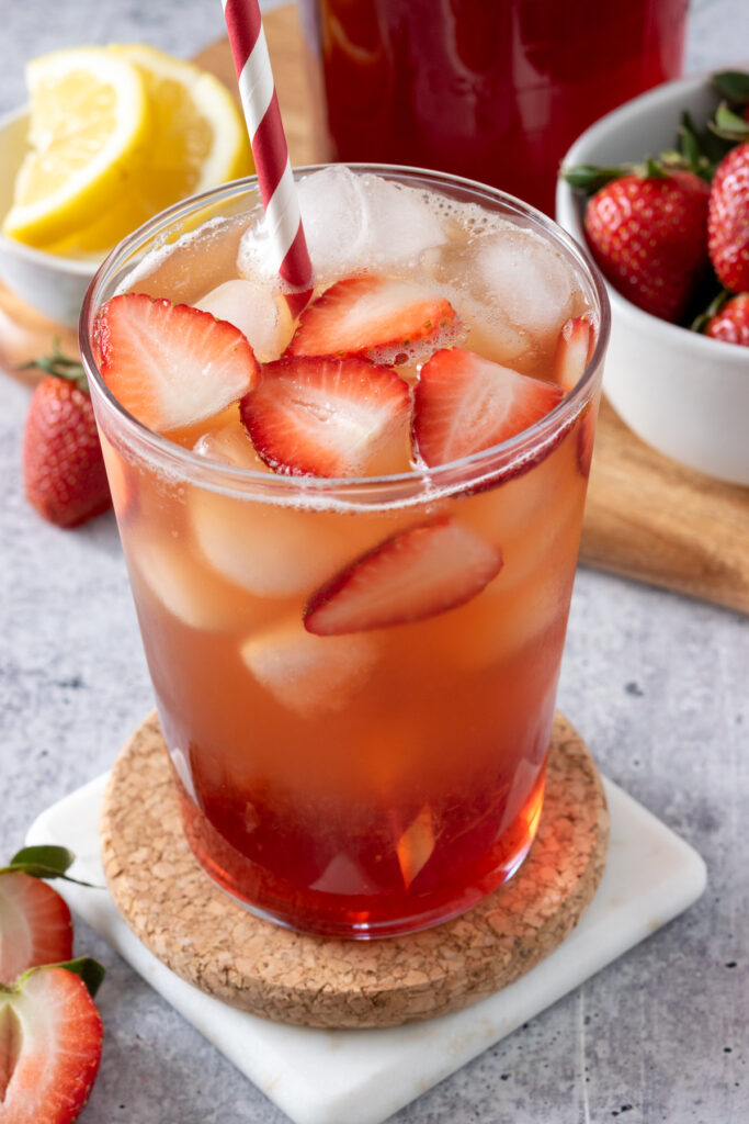 Glass of iced tea with slices of strawberry and ice floating on top, next to a bowl of strawberries and lemons.