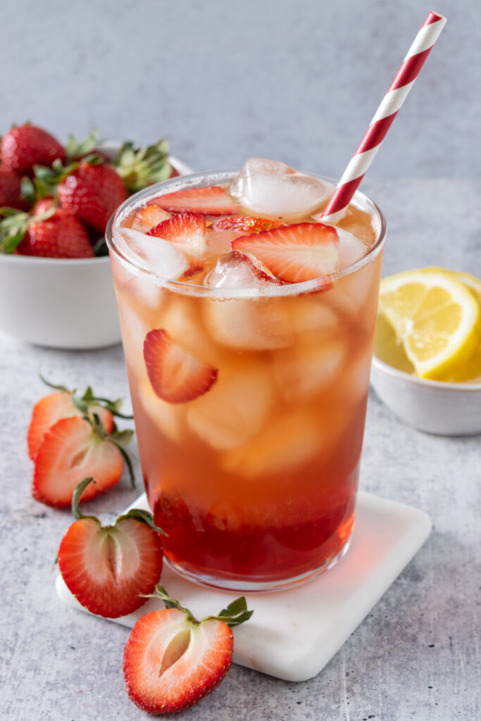 Glass of homemade strawberry iced tea with a red and white striped straw in it and ice and pieces of strawberries floating on top of the refreshing drink.