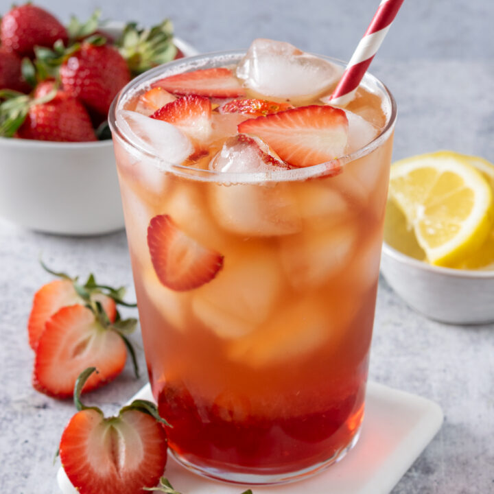 Glass of homemade strawberry iced tea with a red and white striped straw in it and ice and pieces of strawberries floating on top of the refreshing drink.