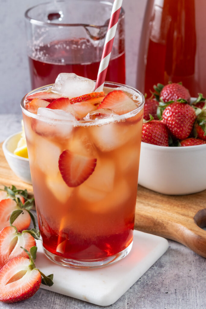 A single serving of strawberry iced tea in a glass on a white coaster, sitting in front of a bottle of homemade strawberry syrup and pitcher of tea.