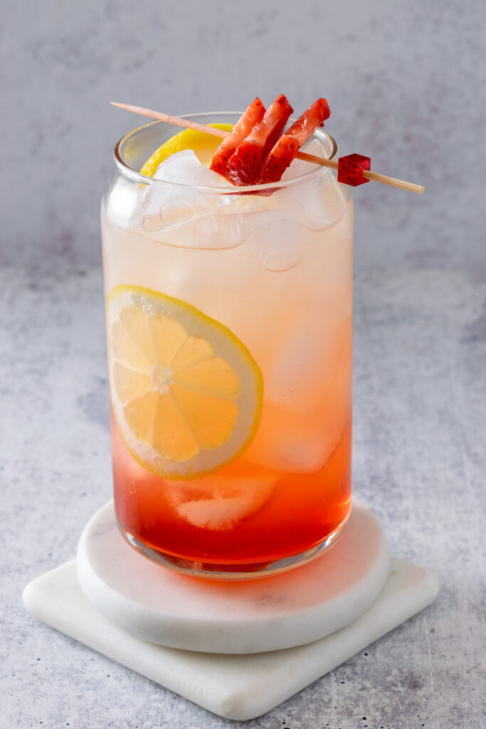 An iced strawberry lemonade that's red on the bottom and has thin slices of lemon in it, and a garnish of sliced strawberries on a toothpick resting across the top of the glass.