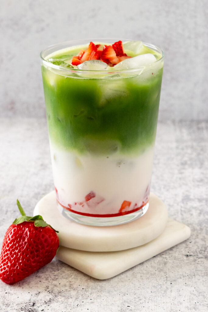 A Strawberry Matcha Latte layered in a glass cup with strawberries on the bottom, followed by ice and milk, matcha and a garnish of chopped strawberries.