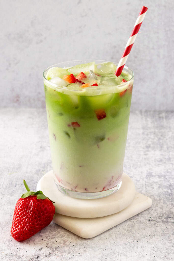 A stirred strawberry matcha latte with bits of real strawberry floating in it, served in a glass with a red striped straw and garnished with more strawberries.