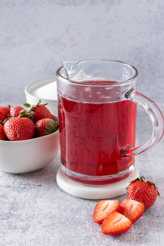 Jar of homemade strawberry simple syrup in front of a bowl of strawberries and a bowl of sugar.