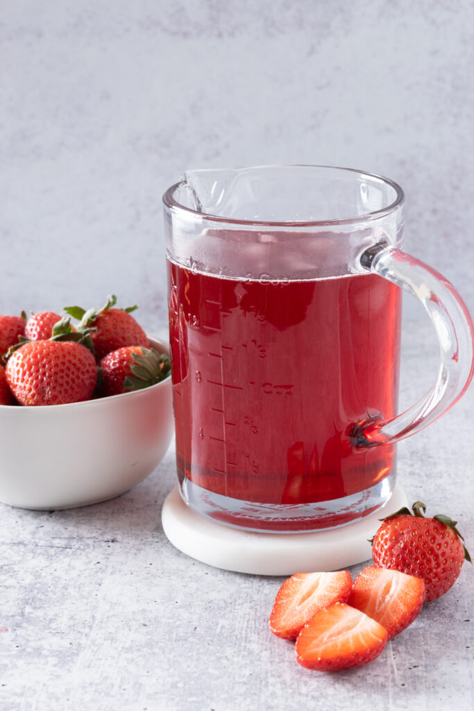 A beautiful jar of bright red strawberry syrup made at home, next to a small white bowl full of strawberries, and three sliced strawberries in front of the jar of syrup.