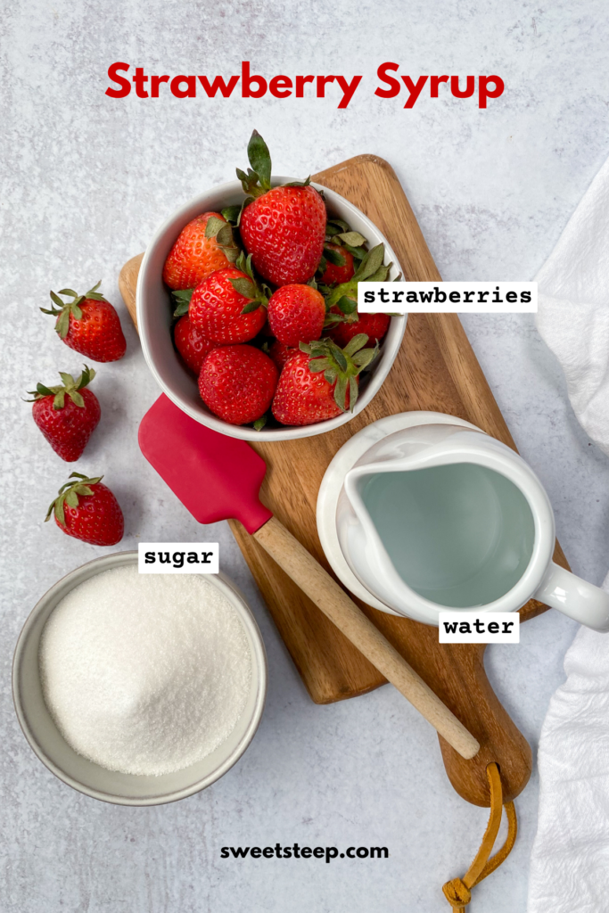 Overhead picture showing a bowl of strawberries and a pitcher of water on a wood cutting board with a red rubber spatula next to them, which is next to a bowl of white sugar and three strawberries.
