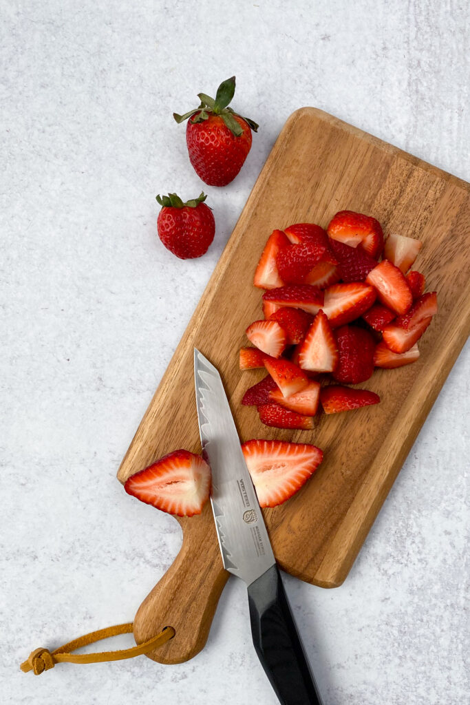 Pile of chopped strawberries and a knife on a wood cutting board.