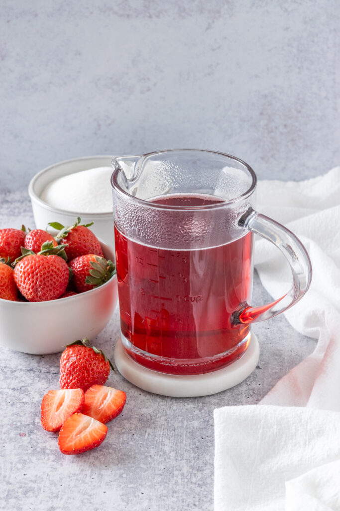 Homemade strawberry simple syrup cooling in a container, next to a bowl of strawberries, bowl of sugar and three sliced strawberries.