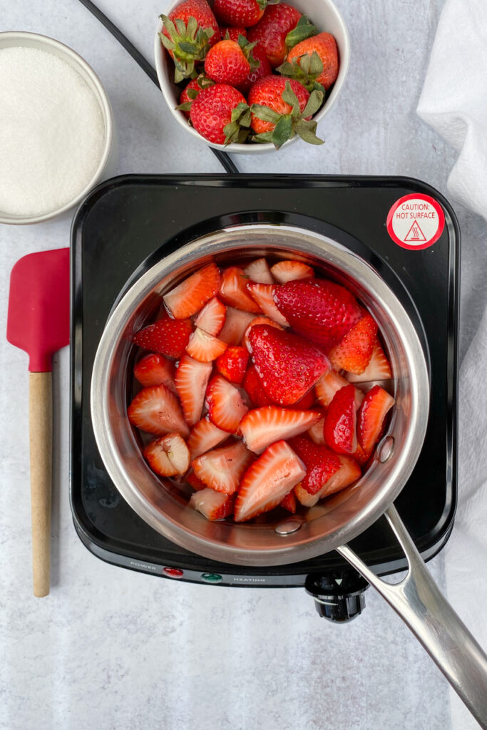 A saucepan on a burner with strawberries and water in it, which is next to a bowl of strawberries, sugar and red rubber spatula.