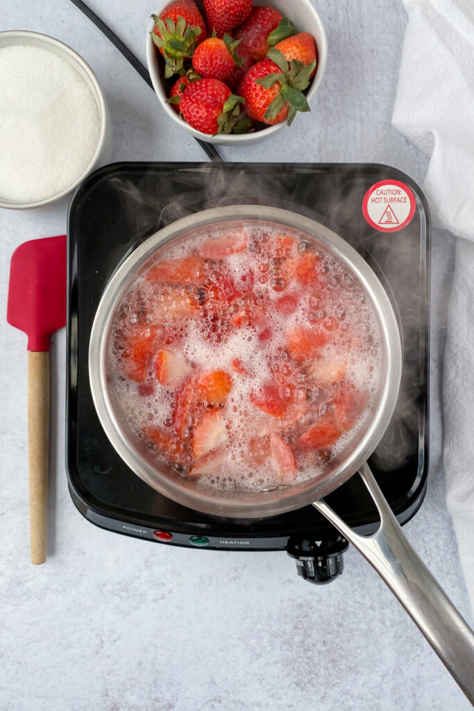 A saucepan on a burner with boiling strawberries and water with lots of bubbles and steam, which is next to a bowl of strawberries, sugar and red rubber spatula.
