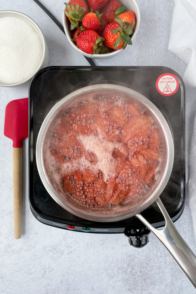 Strawberries and water at a full boil in a saucepan on a burner, showing how the strawberries have turned to a very pale pink color.