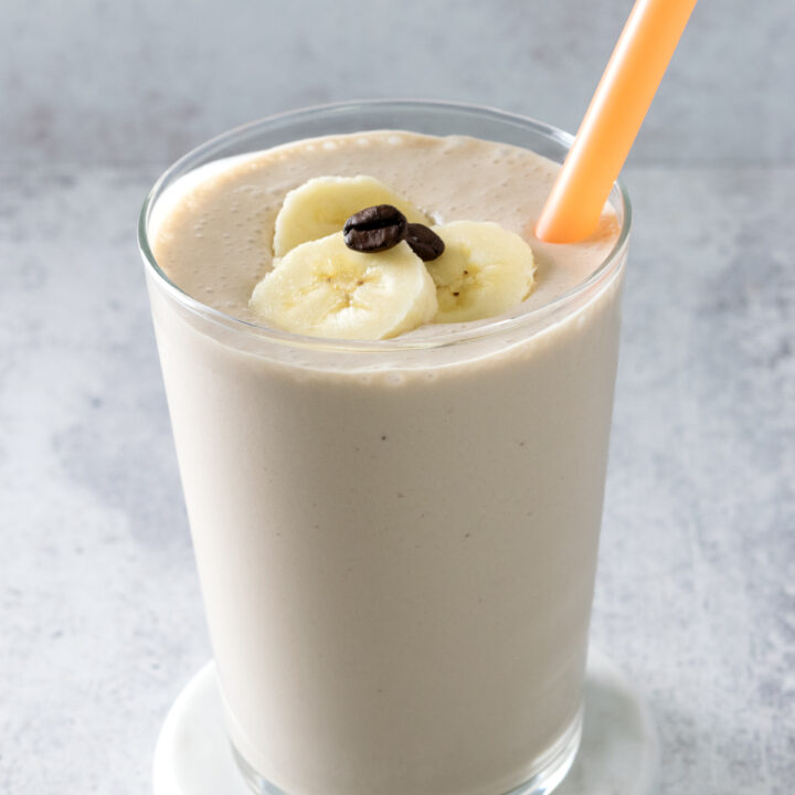 Homemade coffee banana smoothie in a glass with banana slices and coffee beans on top. The smoothie has an orange straw in it.