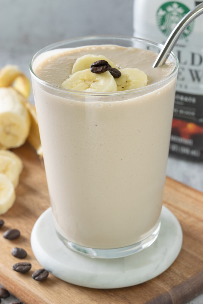A blended coffee banana smoothie in a glass with a straw and three slices of banana on top. The glass is in front of a bottle of Starbucks brand cold brew coffee concentrate and a banana.