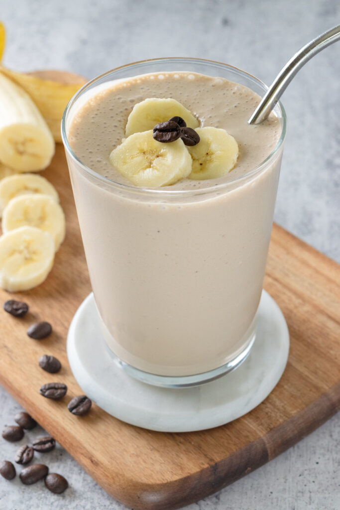A homemade coffee banana smoothie in a glass that's garnished with sliced banana and a few coffee beans. There's a peeled ripe banana and a few more coffee beans next to the smoothie.