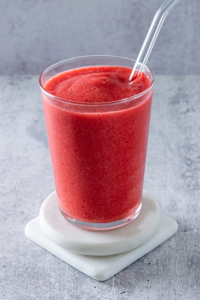 Red-colored frozen raspberry lemonade in glass with a glass straw in it. The drink is sitting on top of two white marble coasters.