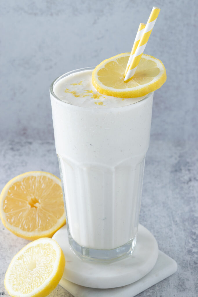 Homemade frosted lemonade beverage in a tall glass cup, which is garnished with a lemon circle and lemon zest. Two yellow and white striped straws are inserted into the lemonade.