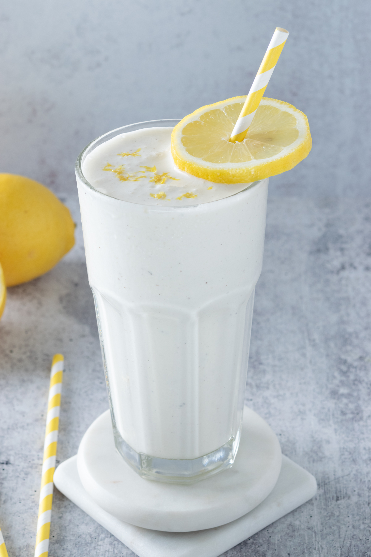 Frosted lemonade drink in a tall frosted glass, garnished with lemon slice and lemon zest. The cup has a yellow striped straw in it and is next to a lemon a a few more straws on the counter.