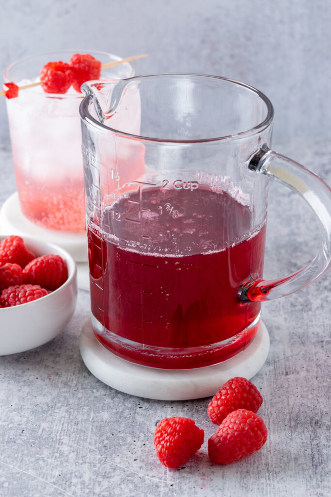 Homemade raspberry syrup in a glass jar next to a small bowl of raspberries and a bubbly pink and white cold drink garnished with fruit.