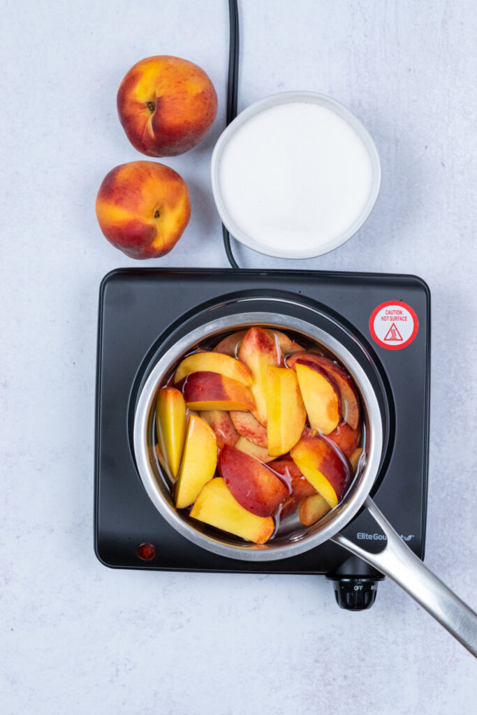 Overhead view of sliced peaches and water in a saucepan on the stove, with whole peaches and a bowl of sugar nearby.