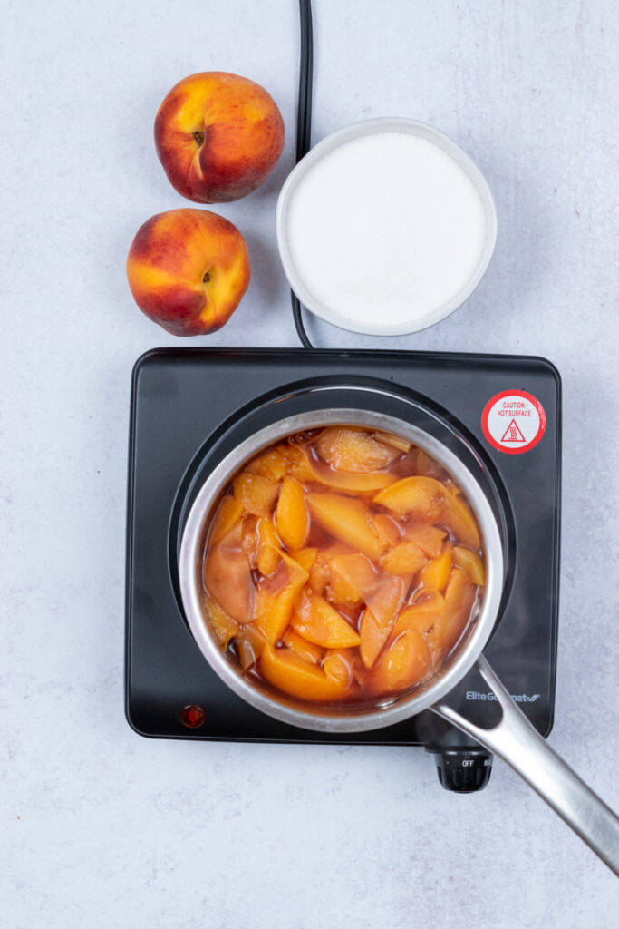 Pale, softened peach pulp in the saucepan after simmering, ready to be strained; nearby are whole peaches and a bowl of sugar.