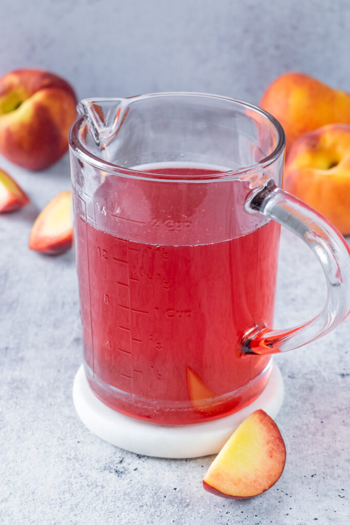 Homemade peach syrup in a glass bottle that's surrounded by sliced and full peaches.