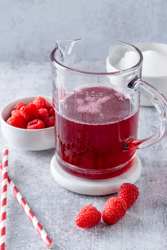 Glass jar half full of raspberry syrup that's a dark pink color. The syrup is next to a bowl of raspberries, bowl of sugar, and two red and white striped straws.