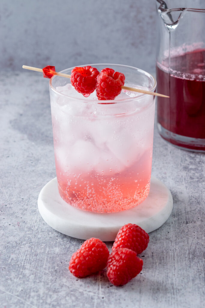 A bubbling glass of sparkling water with layer of raspberry syrup at the bottom. The layered clean and pink iced drink is garnished with two fresh raspberries on a cocktail pick.