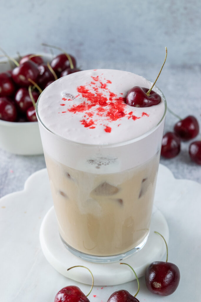 A homemade cherry chai drink in a glass cup that shows the iced chai latte on the bottom and the cherry cream on top. The drink is garnished with crushed cherry candy on top and a fresh cherry with a stem on it.