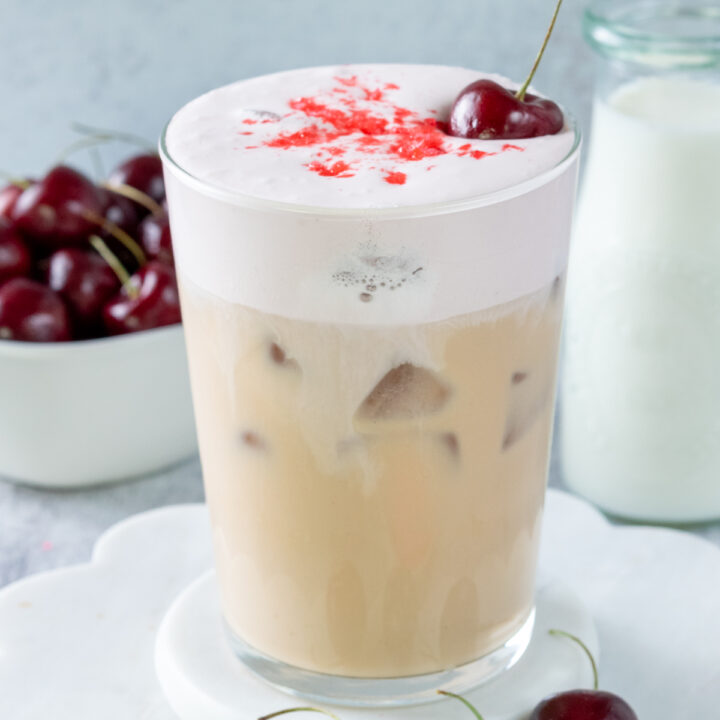 An iced cherry chai in a glass showing the layers in the drink with iced chai on the bottom and a pink colored cherry cream cold foam on top. The drink is garnished with crushed cherry sprinkles, a fresh cherry. In the background is a bowl of cherries and a pitcher of milk.