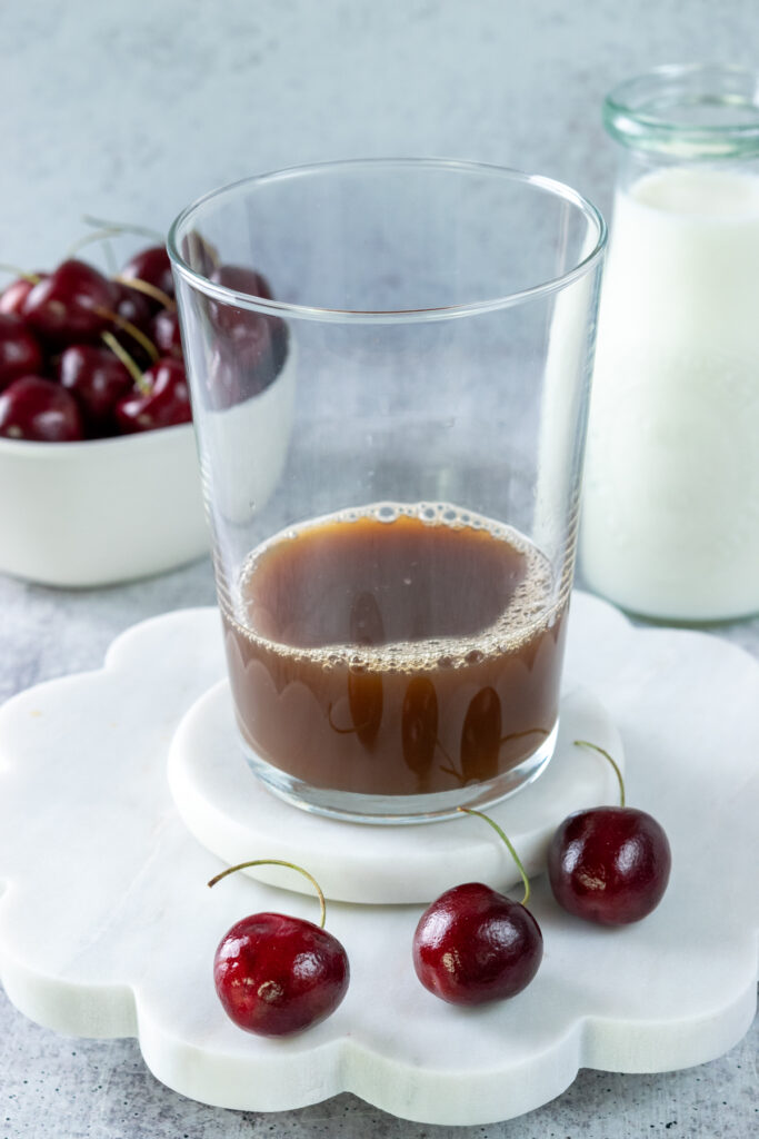 An iced cherry chai is being assembled in a glass cup. The cup has chai concentrate in it. The cup is in front of a bowl of cherries and a pitcher of milk.