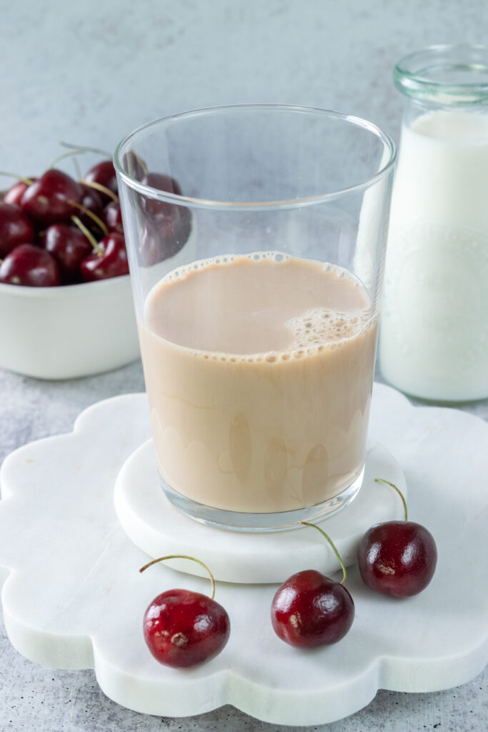 Milk added to chai concentrate in a glass cup that's in front of a bowl of cherries and small pitcher of milk.