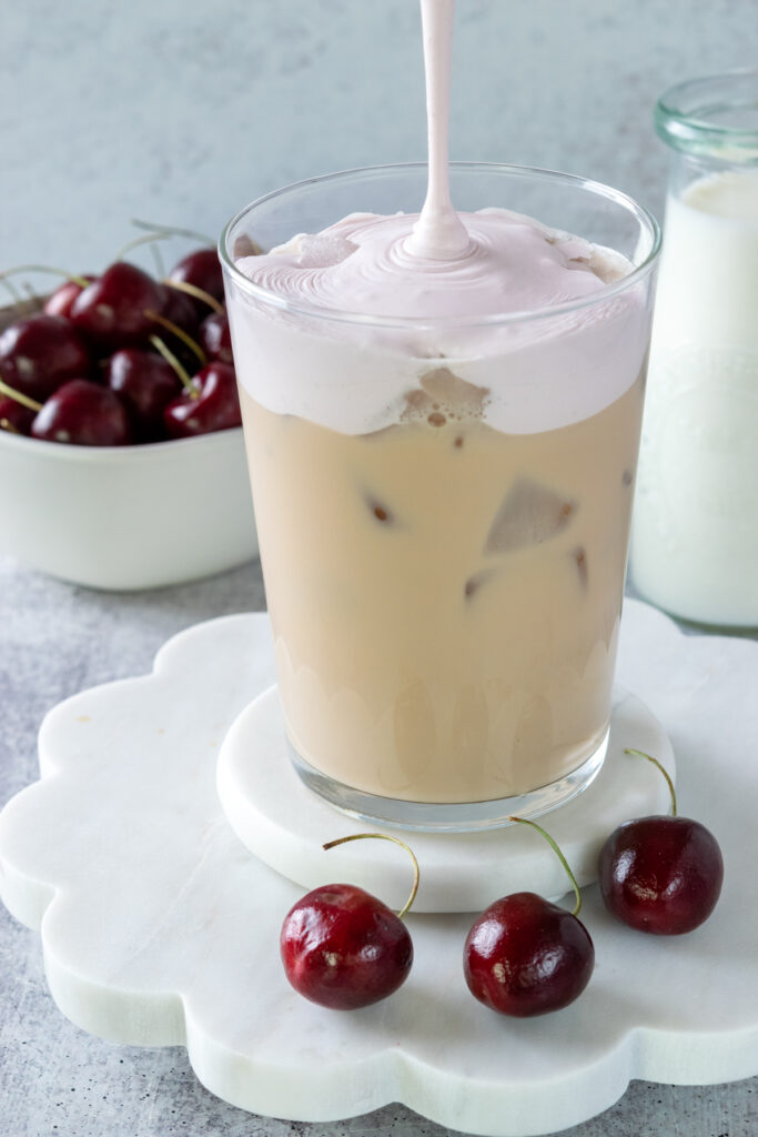 A thick stream of cherry cream cold foam being poured in a pink-colored layer on top of the iced chai drink.