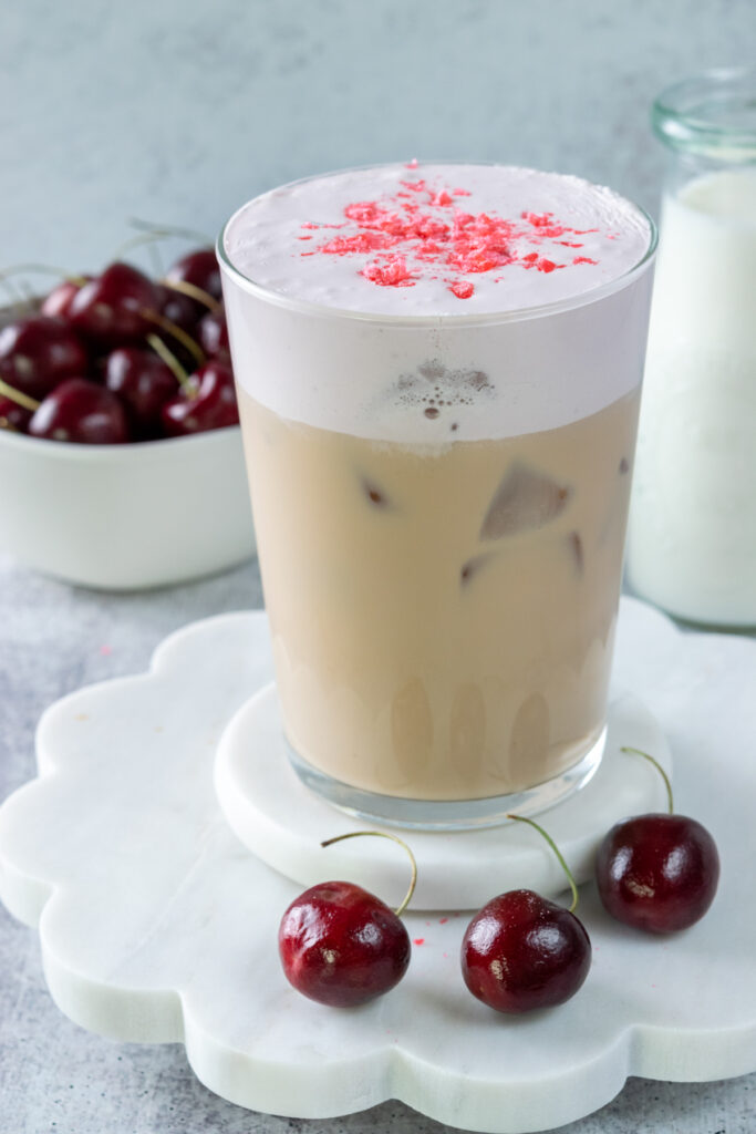A homemade iced cherry chai garnished with crushed cherry candy on top of the cherry cream cold foam. The layered drink is tan on the bottom with a thick layer of pink cherry cream.