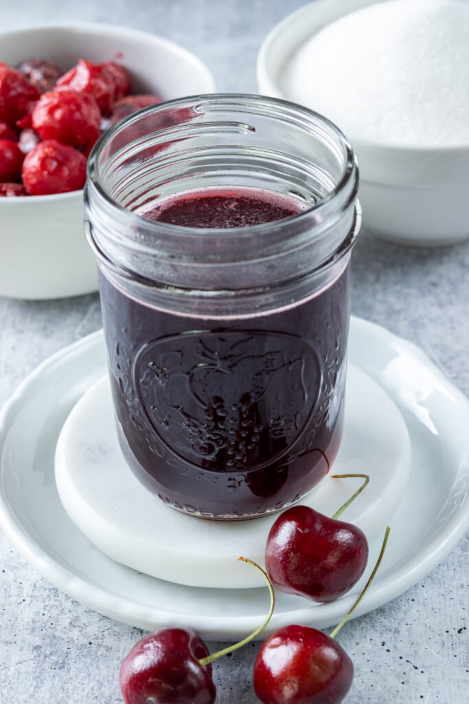 Close-up view of cherry syrup in a glass jar showing its deep red color with some pinkish bubbles around the edges. There's a bowl of frozen cherries and bowl of sugar behind the jar of homemade simple syrup.